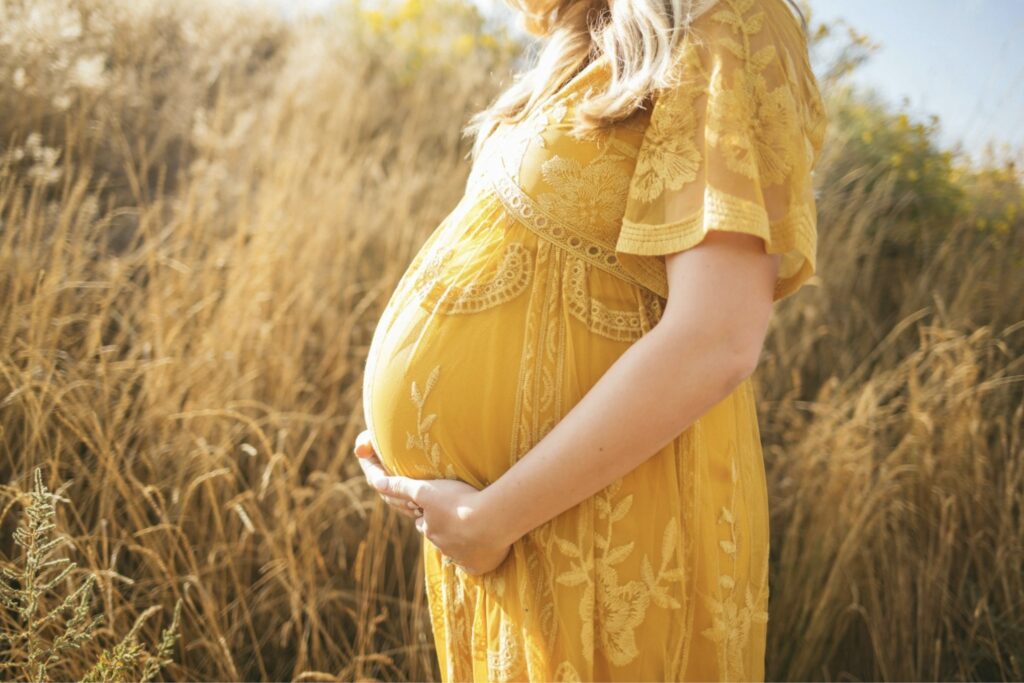 A pregnant woman in a yellow lace dress stands in a sunlit field, gently holding her baby bump surrounded by tall golden grass.