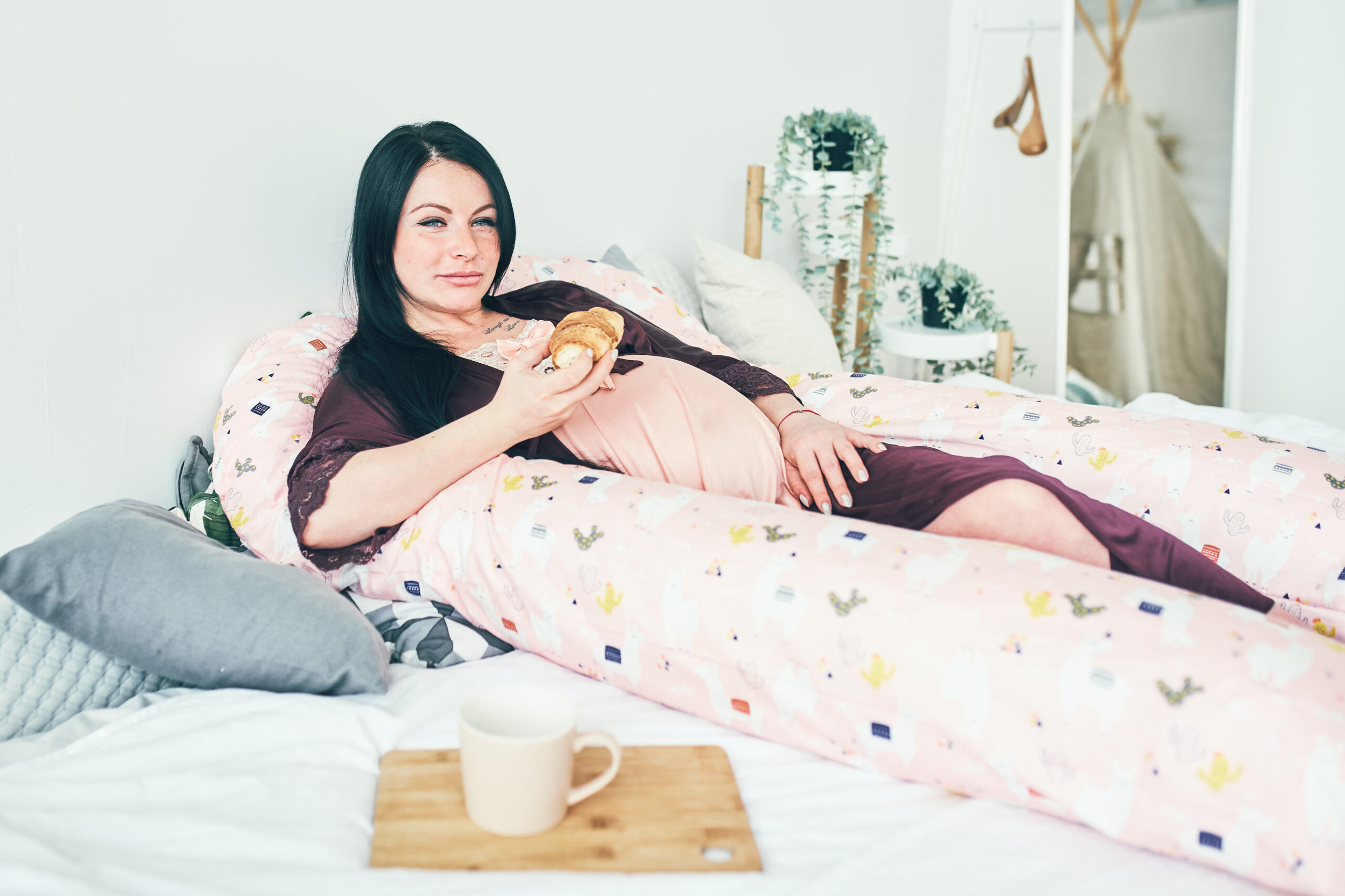 A pregnant girl with dark hair is lying in bed on a maternity pillow, drinking tea and eating croissants. The happiness of waiting for a child.